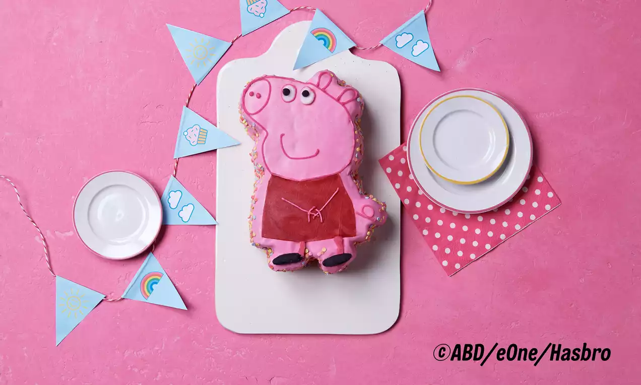 Peppa-Wutz-geformter Kuchen mit rosa Zuckerguss und Details in Rot, Schwarz und Weiß auf weißer Platte, dekoriert mit bunten Wimpeln und Geschirr
