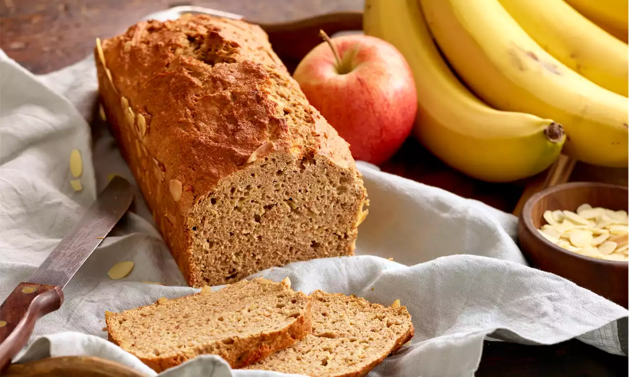 Goldbraunes Banana Bread mit lockerer Textur auf hellgrauem Stofftuch, daneben frische Bananen, roter Apfel und Mandelblättchen in Schale