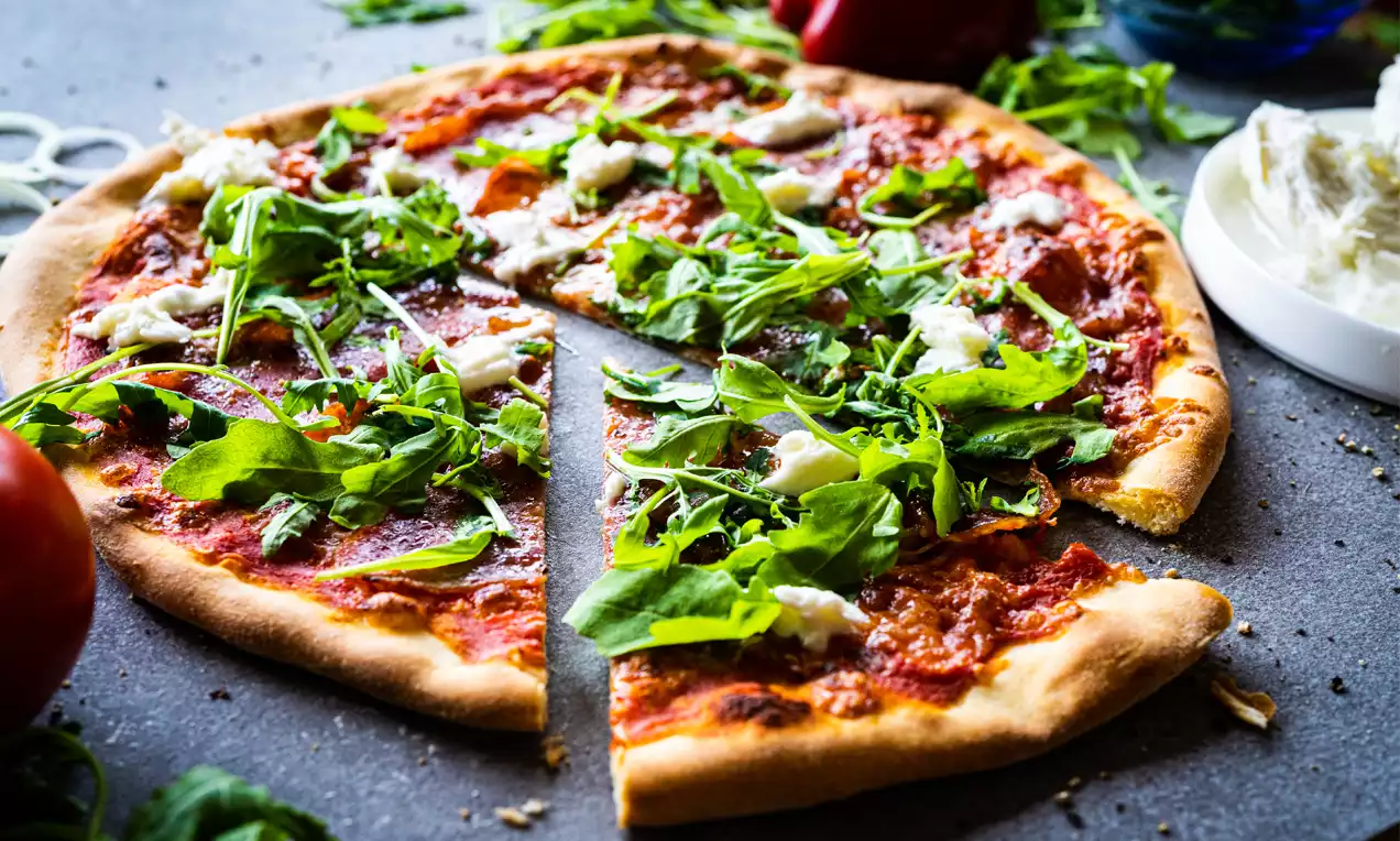 Pizza à croûte dorée garnie de roquette vive, fromage fondant et sauce rouge riche sur une surface sombre avec des légumes frais