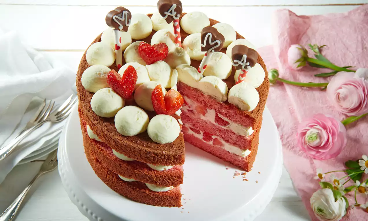 Rosafarbene Torte mit hellen Sahnetupfen, frischen Erdbeerherzen und Schokoladen-Dekoren auf einer weißen Tortenplatte, umrahmt von Blumen