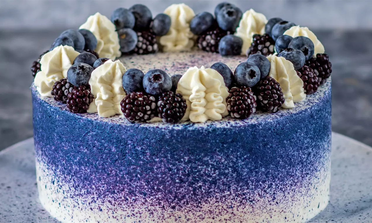 Кръгла торта с преливащи нюанси от бяло към наситено лилаво, украсена с венец от къпини, боровинки и кремови розетки върху сива чиния
