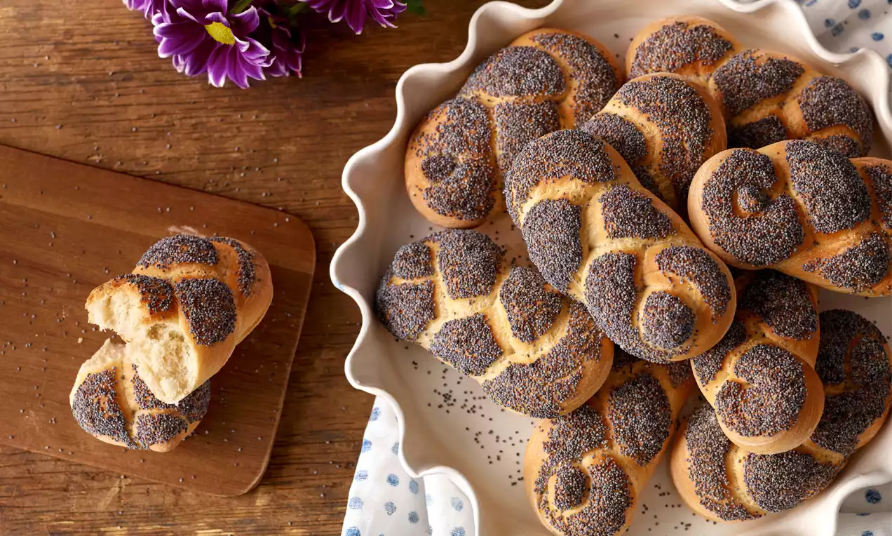 Kleine, goldbraune, geflochtene Mohnweckerl mit dunkler Mohnkruste in einer weißen Keramikschale auf Holztisch mit Holzbrett