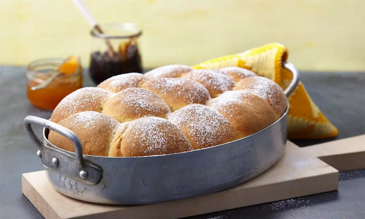 Златисти Великденски кифлички с пухкава текстура, поръсени с фин слой пудра захар, подредени плътно в овална метална тава на светъл фон