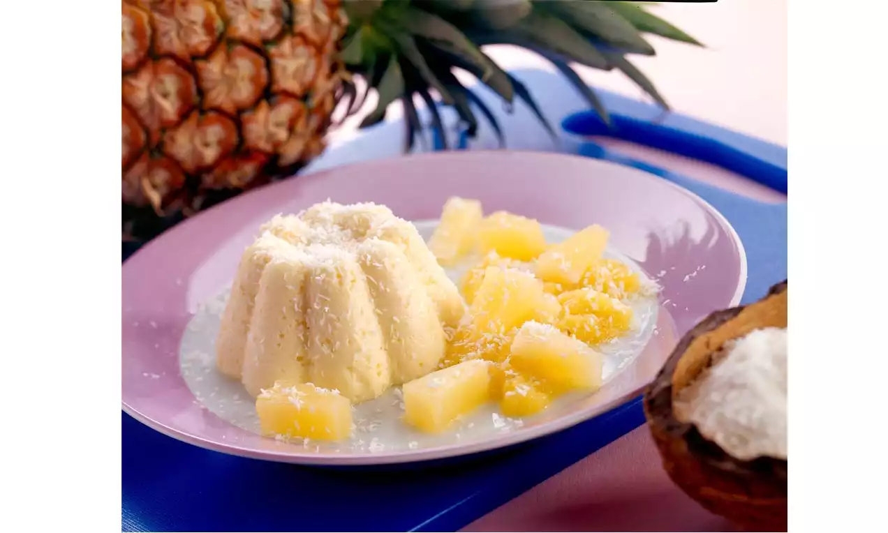 Mousse jaune pâle en forme de fleur entourée de dés d’ananas juteux, saupoudrée de noix de coco sur une assiette rose