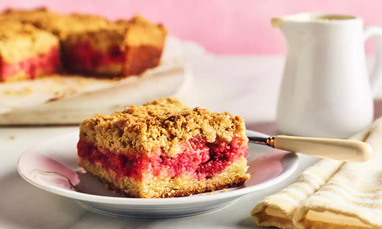 Golden crumbly topping on a square slice of rhubarb crumble cake with vibrant red rhubarb layer on a white plate beside a cream jug