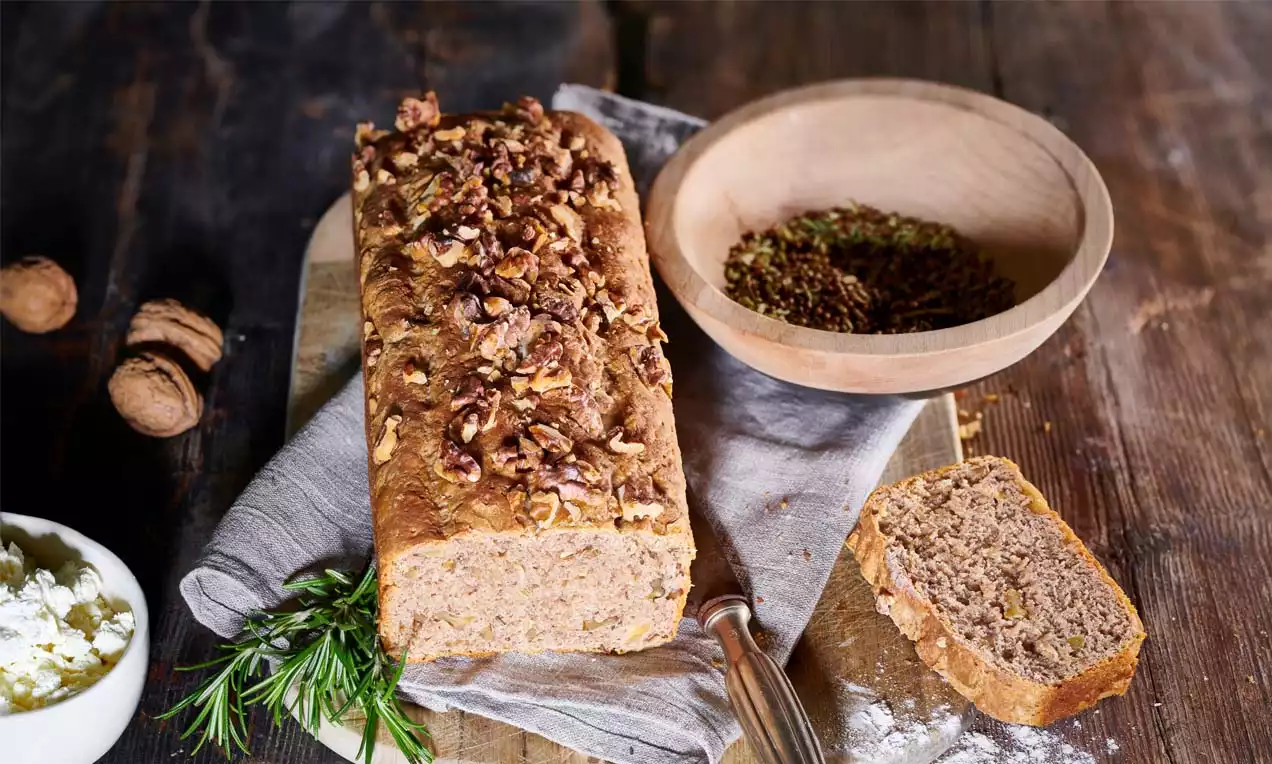 Goldbraunes Dinkel-Walnussbrot mit groben Nussstücken auf einem Leinentuch, daneben ein Holzmesser, Rosmarinzweige und Leinsamen in einer Holzschale