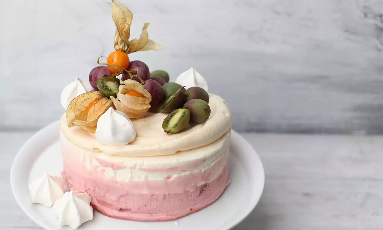 Vacherin Glacé-Torte mit rosafarbenen und weißen Schichten, gekrönt von Physalis, Trauben, Kiwi und Baisers auf weißem Teller