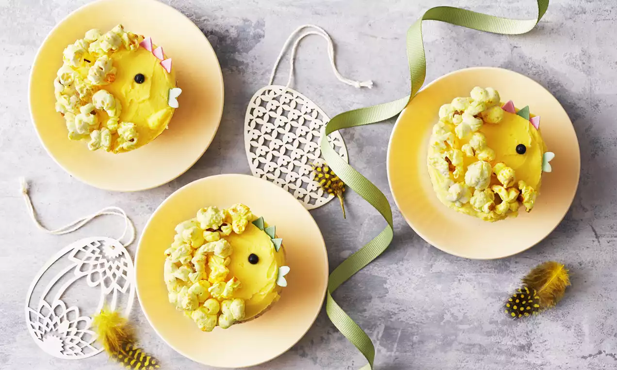 Påskegule muffins formet som kyllinger med krusede popcornkanter, sorte sukkerøjne og farvede papirkamme på lyse tallerkener med bånd og pynt