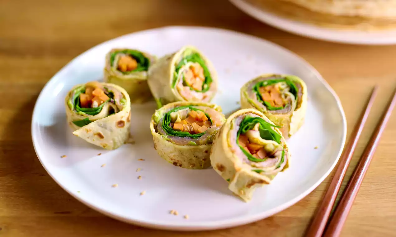 Roulés de crêpes dorées en spirale garnis de légumes frais colorés et de saumon, disposés sur une assiette blanche avec des baguettes