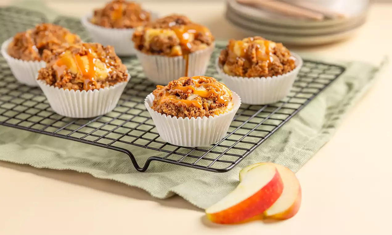 Appelmuffins met een kruimelige speculaaslaag en goudkleurige karameldrizzle, gerangschikt op een zwarte taartrooster met appelpartjes ernaast