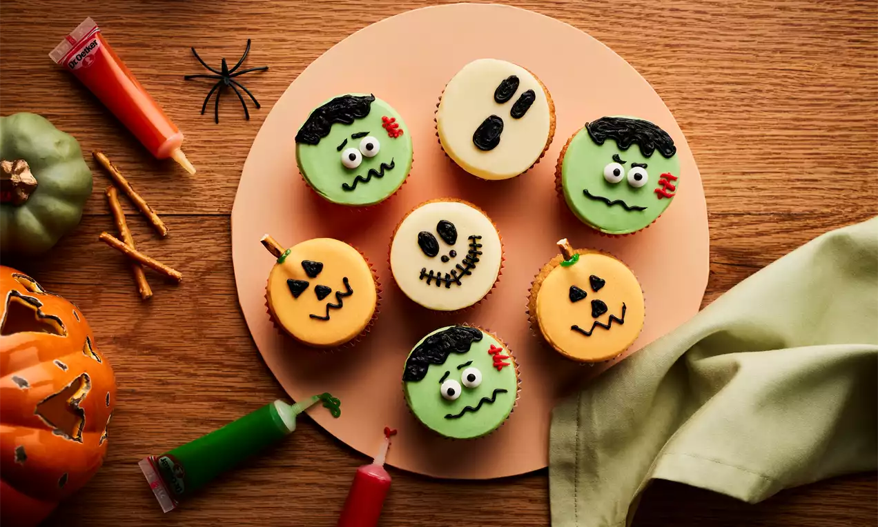 Halloween cupcakes with green Frankenstein faces, orange pumpkin designs, and white skulls arranged on a round beige plate with Halloween-themed decorations nearby