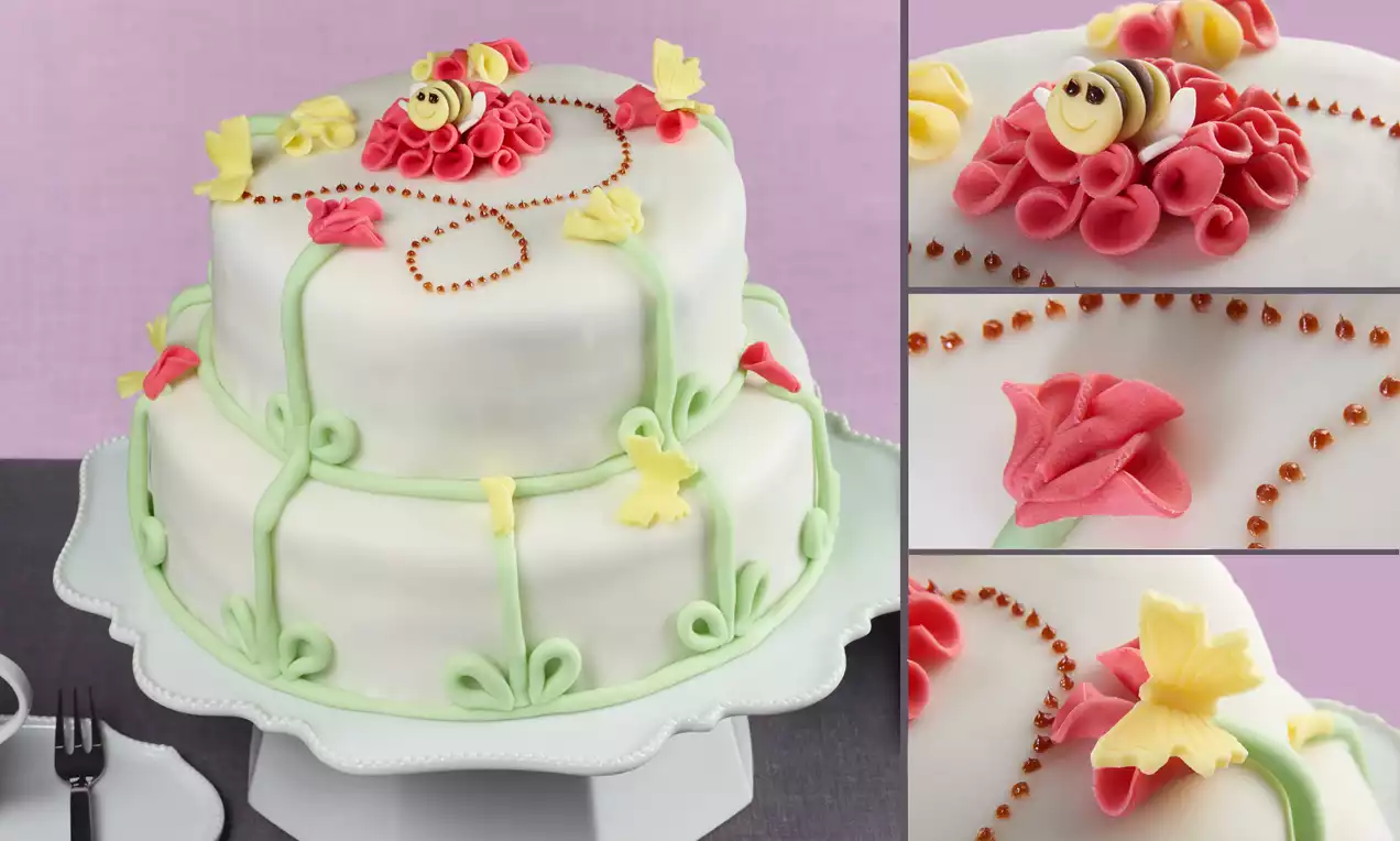 Weiße Fondant-Torte mit grünen Ranken, bunten Blüten und einer gelben Bienen-Figur, dekorativ auf einer weißen Tortenplatte präsentiert