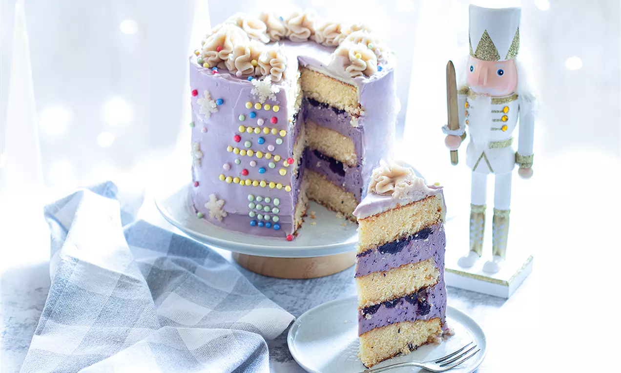 Висока кръгла торта с лилава глазура, цветни захарни декорации и кремови рози, нарязана на пластове с крем и плодов пълнеж