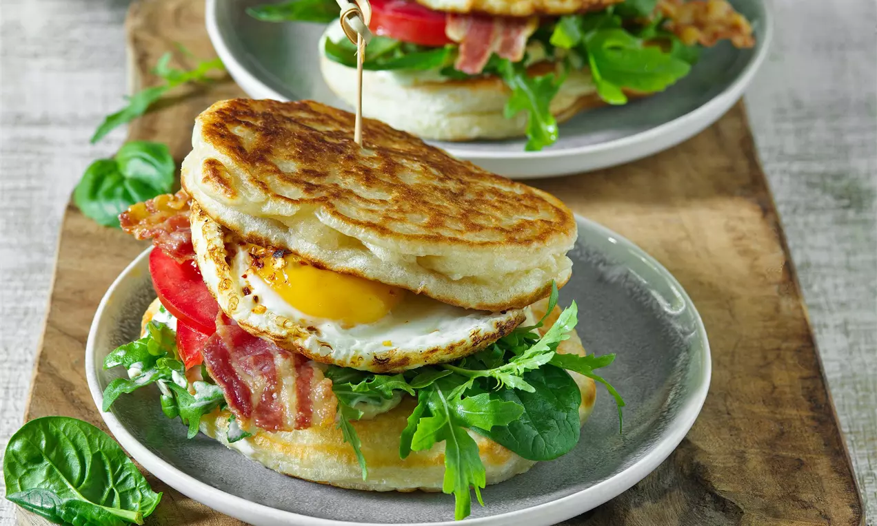 Sendvič z nadýchaných lívanců se zlatavou kůrkou, vrstvený s čerstvými listy zeleniny, plátky rajčat, křupavou slaninou a vejcem na bílé misce