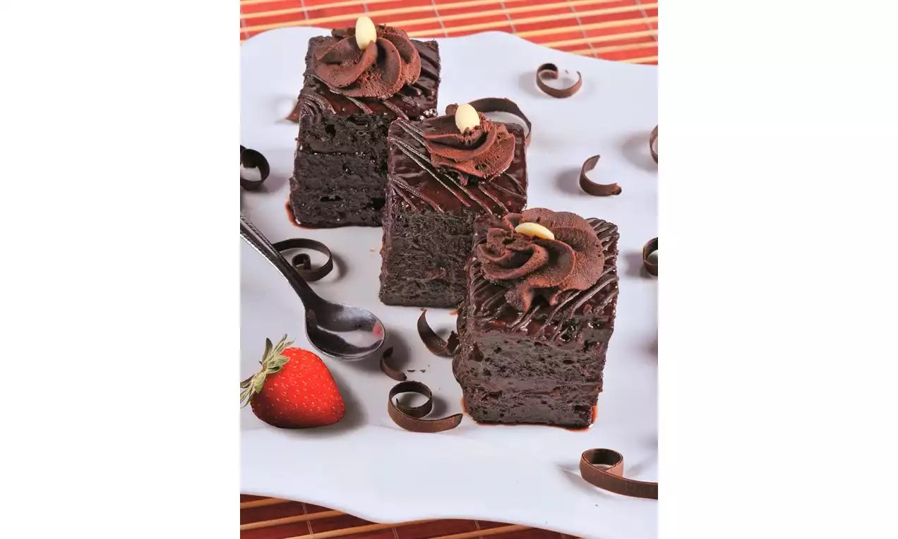 Prăjituri Amandina de post cu straturi de blat umed ciocolatiu, decorate cu cremă densă și glazură, alături de o căpșună și spirale de ciocolată