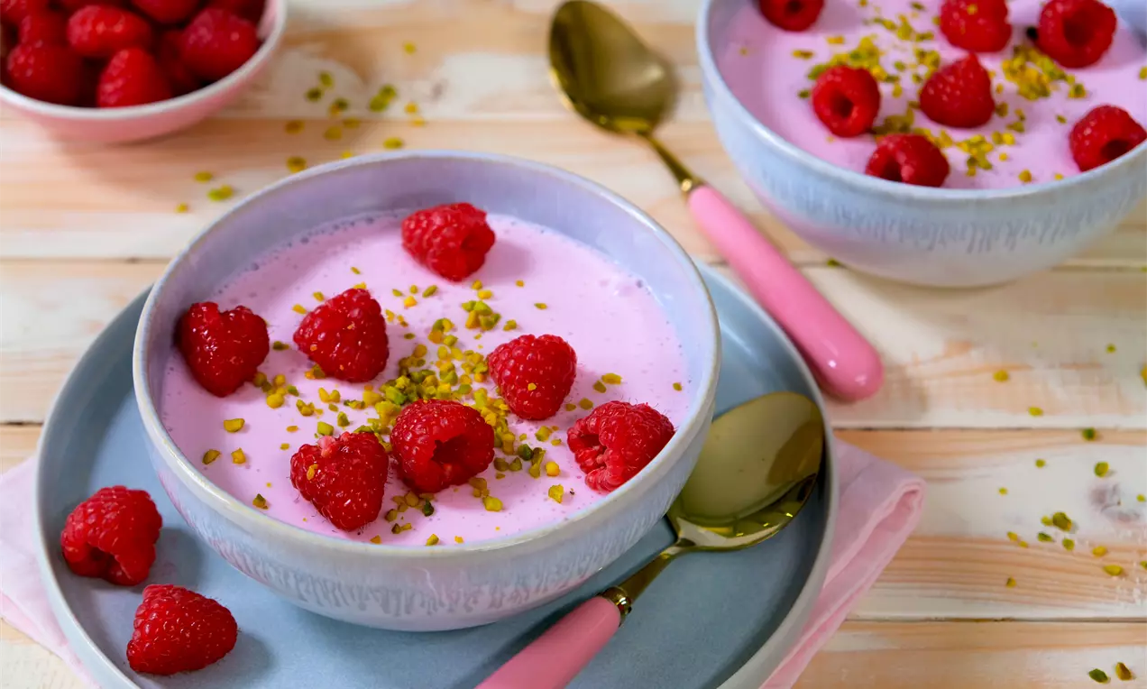 Rosa Quarkcreme in einer Keramikschale, mit frischen Himbeeren und gehackten Pistazien garniert, auf einem Teller mit rosa Besteck