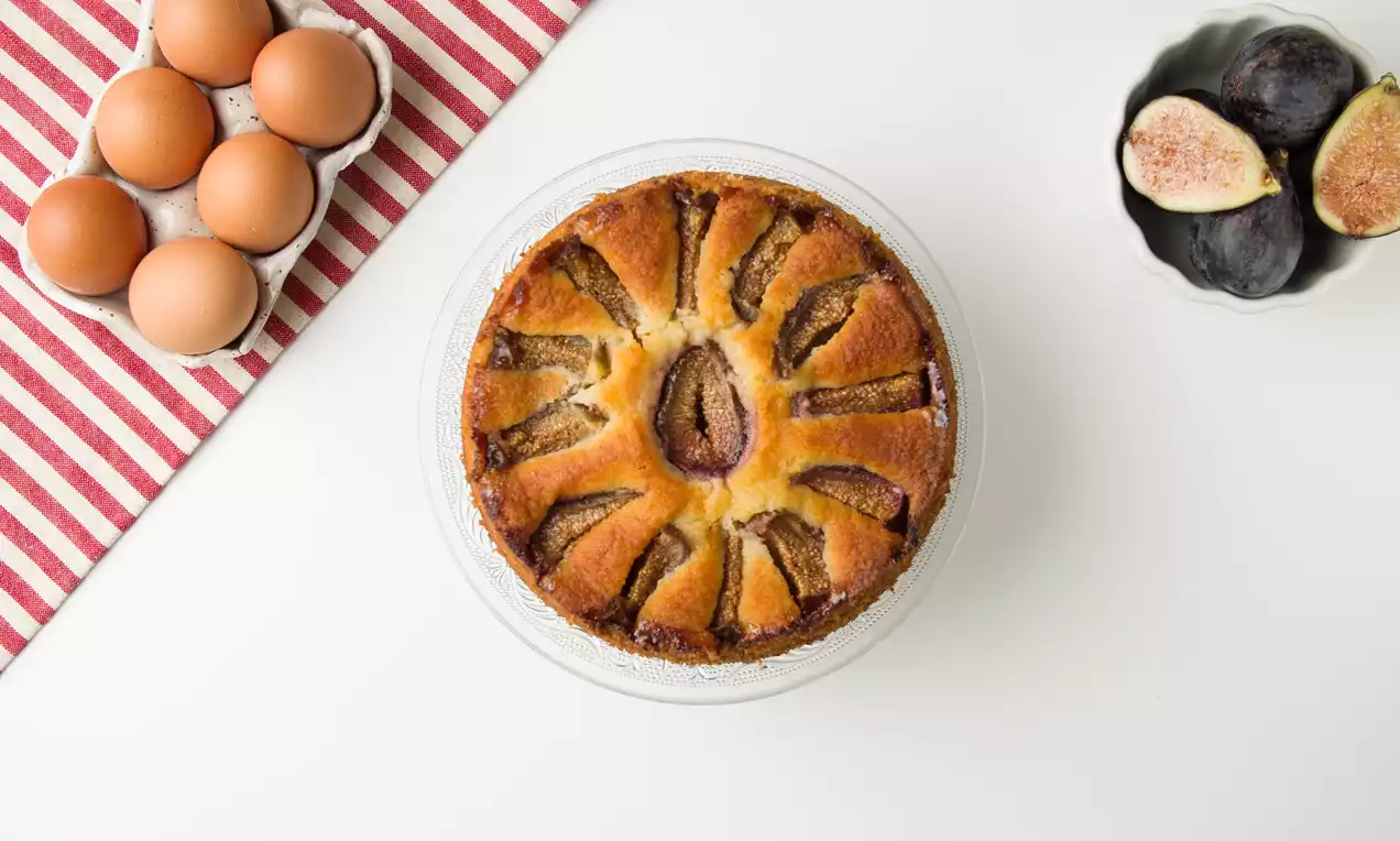 Tarta dorada con bordes ligeramente tostados, higos abiertos en abanico sobre la superficie y almendras en un plato de cristal