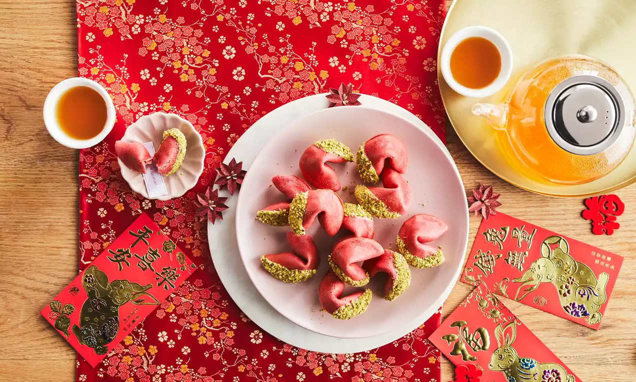 Pink fortune cookies with crescent shapes, dipped in crushed green pistachios, arranged on a white plate on a red floral cloth