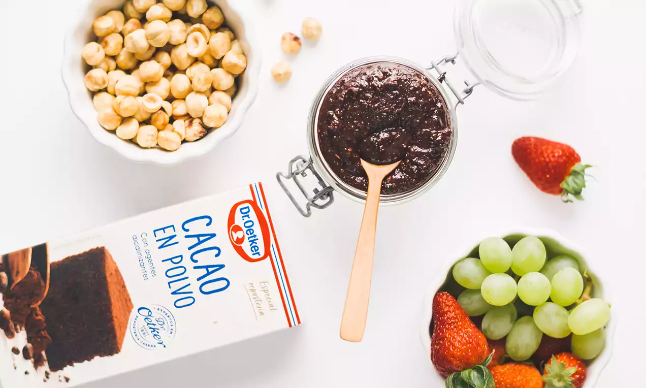 Crema espesa de color marrón oscuro con textura lisa, dispuesta en un tarro de cristal abierto junto a frutas, avellanas y cacao en polvo