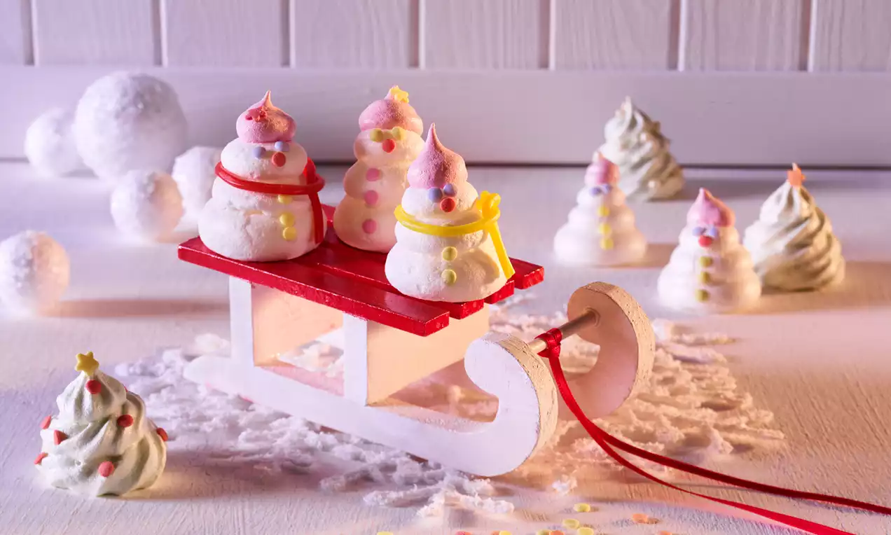 Små hvite marengs formet som snømenn med rosa hatter og som juletrær med stjerner og strø pyntet på en rød kjelke og hvit bakgrunn