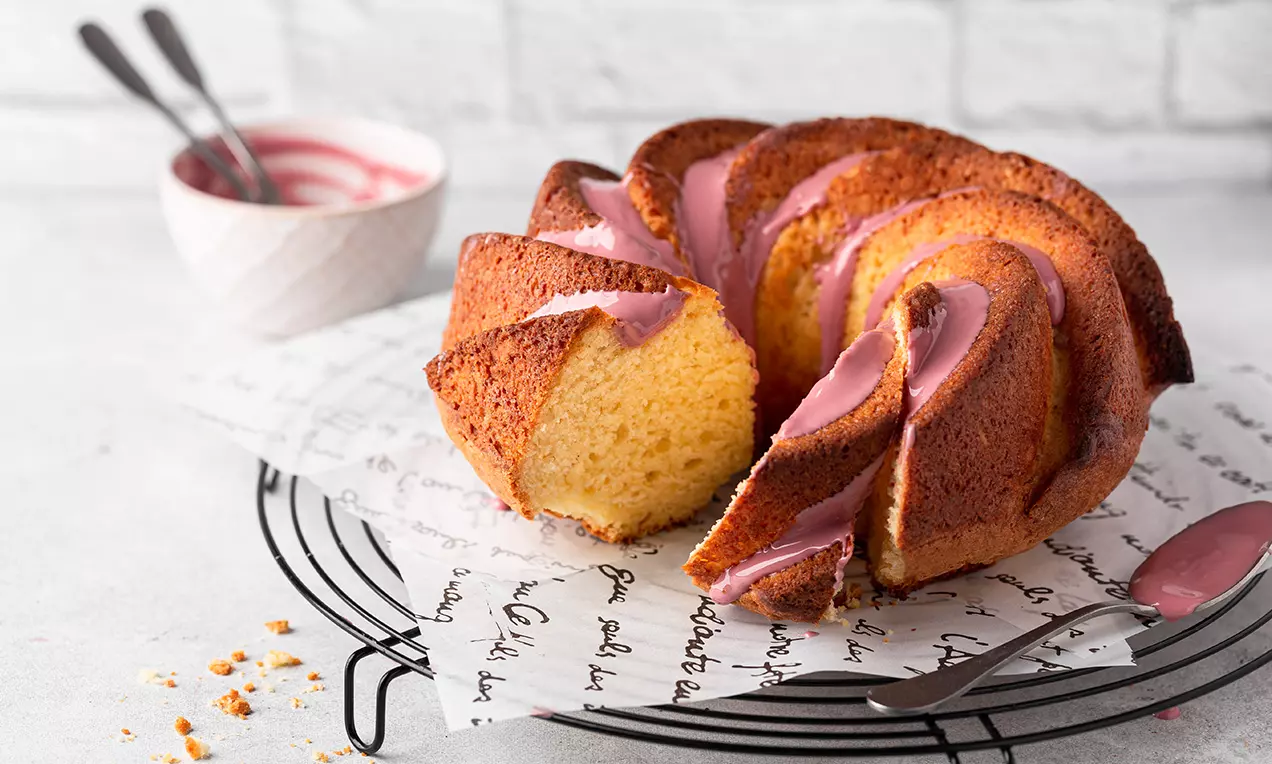 Saftiger, goldbrauner Gugelhupf mit rosa Glasur auf einem schwarzen Kuchengitter, begleitet von einer Schale Glasurcreme und einem Löffel