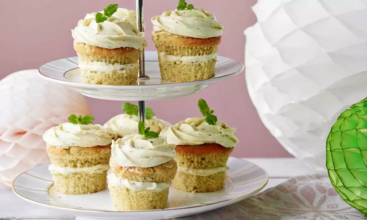 Mojito-Cupcakes mit weißlich-grüner Limettencreme und Minzblättern auf einer zweistufigen Etagere vor rosa und grüner Dekoration