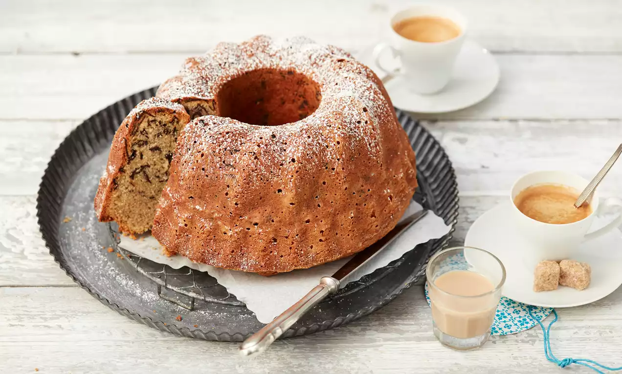 Baileys®-Nuss-Gugelhupf, goldbraun mit Walnussstücken, heller Sahneglasur und dunklen Schokoladenraspeln auf weißem Teller angerichtet