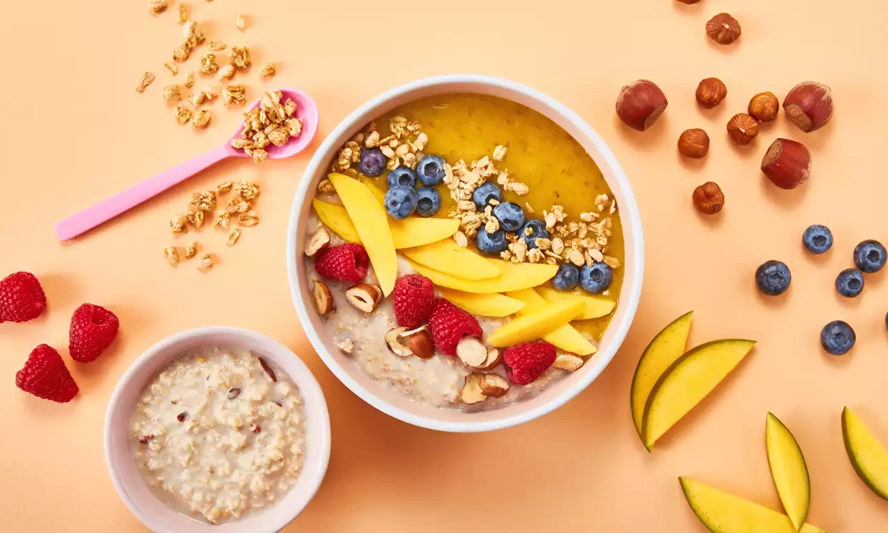 Bunte Frühstücksbowl mit gelber Mango, roten Himbeeren, blauen Heidelbeeren, Haselnüssen und Granola dekorativ auf cremiger Basis