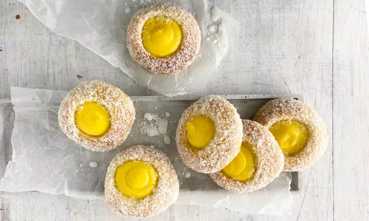 Skoleboller dorati e soffici con centro di crema gialla lucida, bordi cosparsi di cocco bianco, disposti su carta cerata sopra un tavolo chiaro