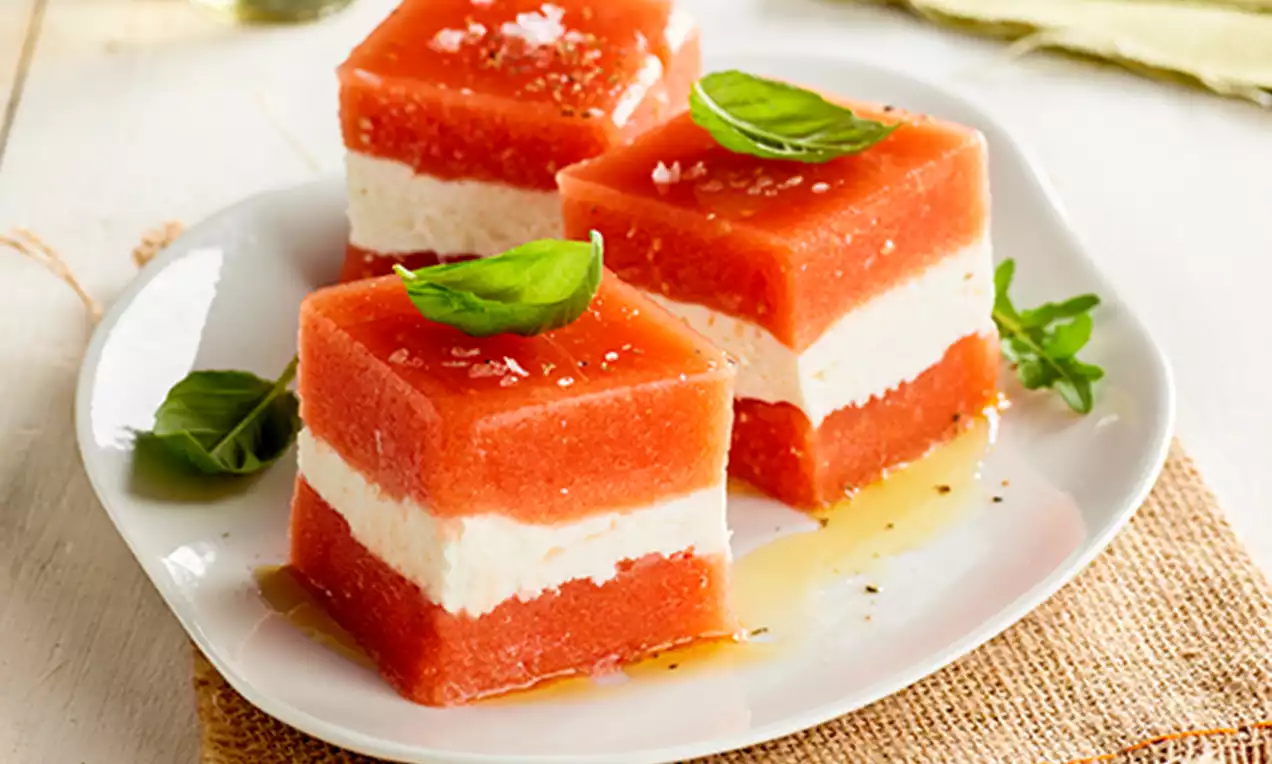 Cubos rojos y blancos de sandía y queso crema en capas, decorados con hojas de albahaca y sal sobre un plato blanco con miel