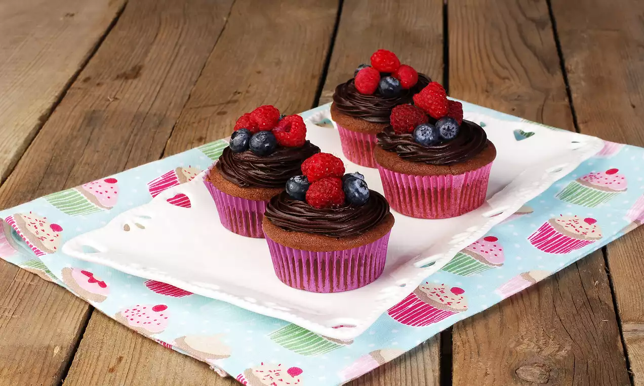 Mor kağıtlara sarılmış, kremalı üstleri orman meyveli süslenmiş dört muffin, desenli örtü üzerinde beyaz tabakta sergilenmiş