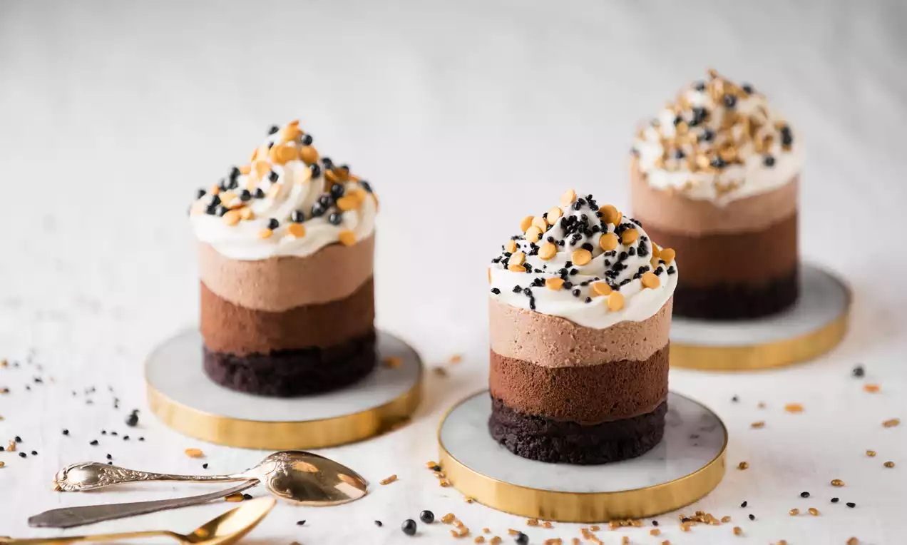 Små sjokoladekaker i lagdelte nyanser, toppet med krem, svarte og oransje sukkerkuler, på marmorfat med gullkant og bestikk ved siden av