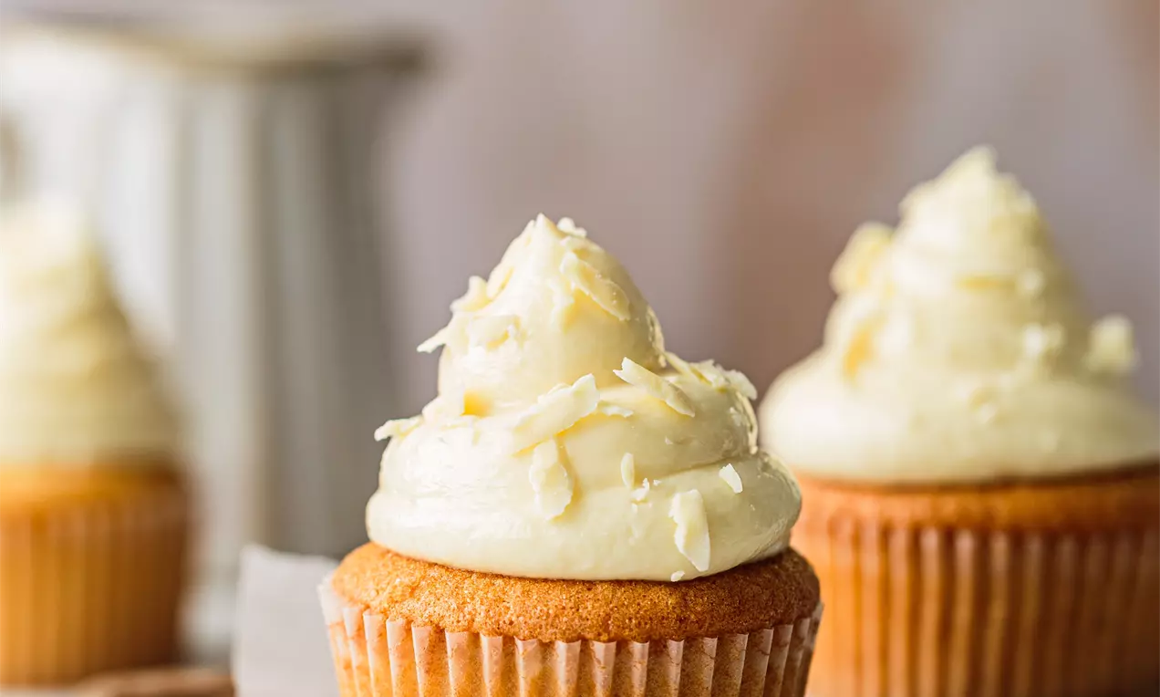 Picture - Vanilla cupcakes with golden sponge bases and swirls of pale cream frosting topped with white chocolate flakes on a wooden board