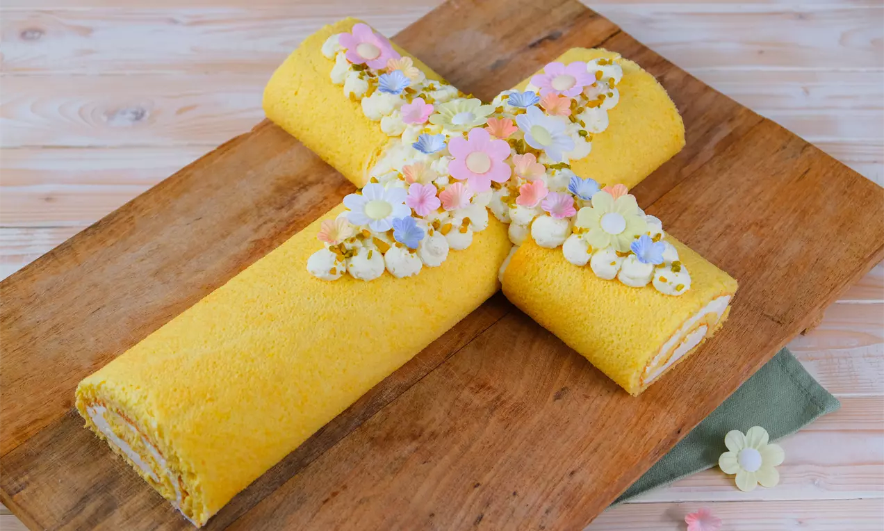 Gelbe Biskuitrolle in Kreuzform mit heller Creme und bunten Blüten dekoriert, auf rustikalem Holzbrett mit pastellfarbenen Akzenten drapiert