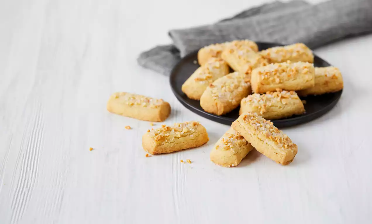 Rektangulära finska pinnar gyllenbruna med sockerkant och hackad nötströssel på svart tallrik mot vit träbakgrund med grå duk