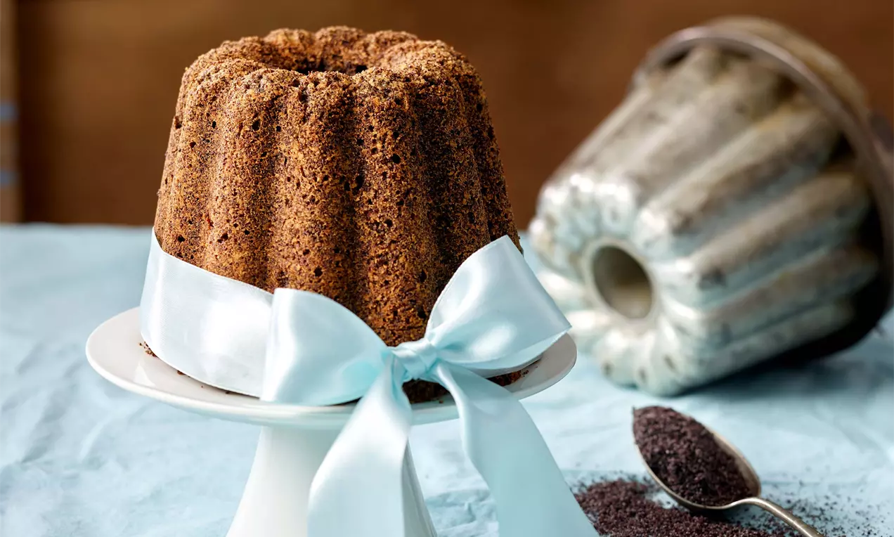 Maková bábovka s tmavě hnědou křupavou krustou na bílém podnosu zdobená světle modrou stuhou, vedle lžíce s mákem a kovová forma