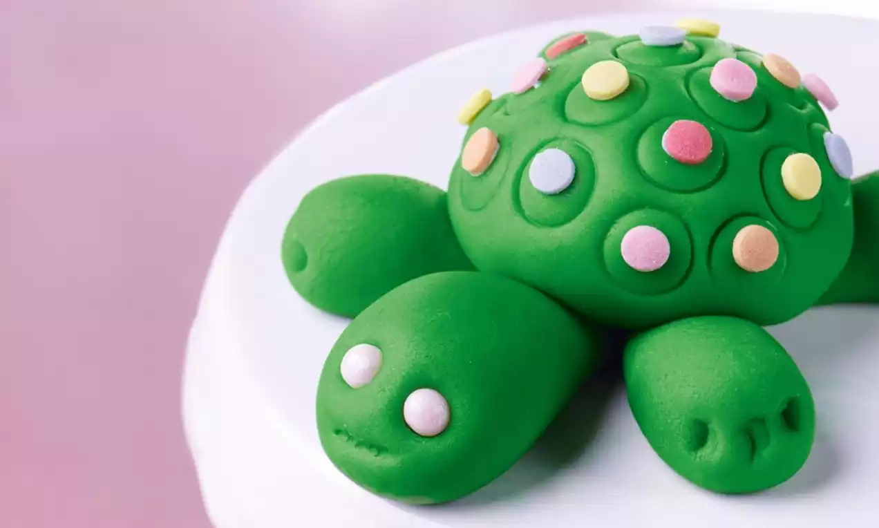 Zielony żółw z masy cukrowej o gładkiej fakturze, ozdobiony kolorowymi cukrowymi kropkami, leżący na białej powierzchni