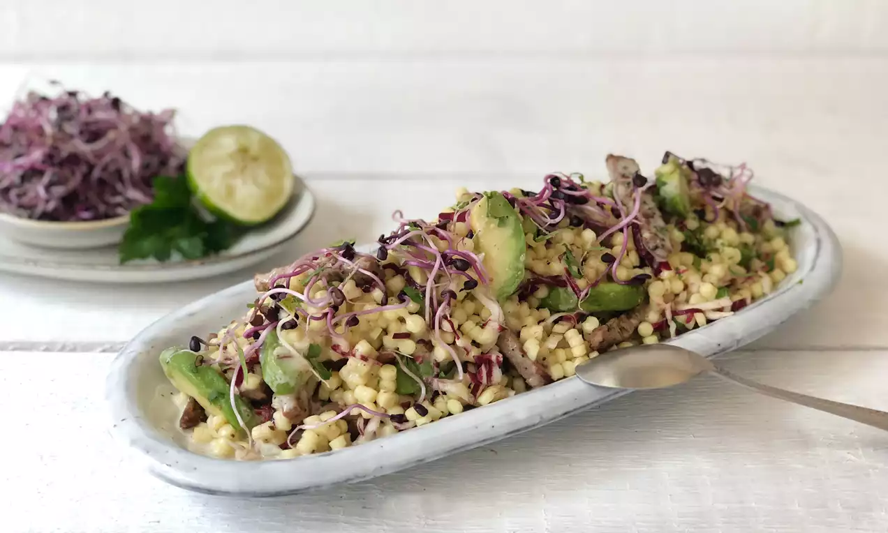 Fregola-Salat mit Avocado-Scheiben und lila Sprossen auf länglichem weißen Teller, daneben Limettenscheibe und Kräuter auf kleiner Platte