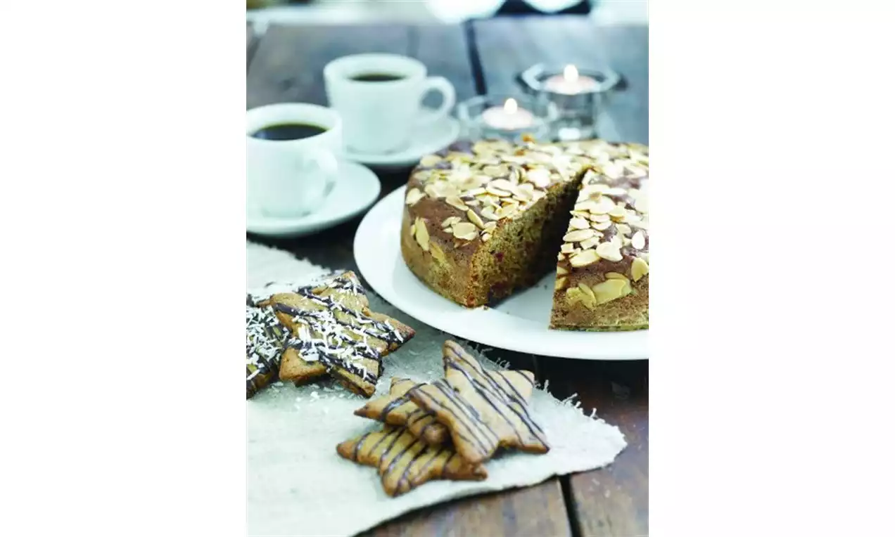 Saftig krydderkake pyntet med skivede mandler på et hvitt fat sammen med småkaker på linserviett med to kaffekopper og telys i bakgrunnen