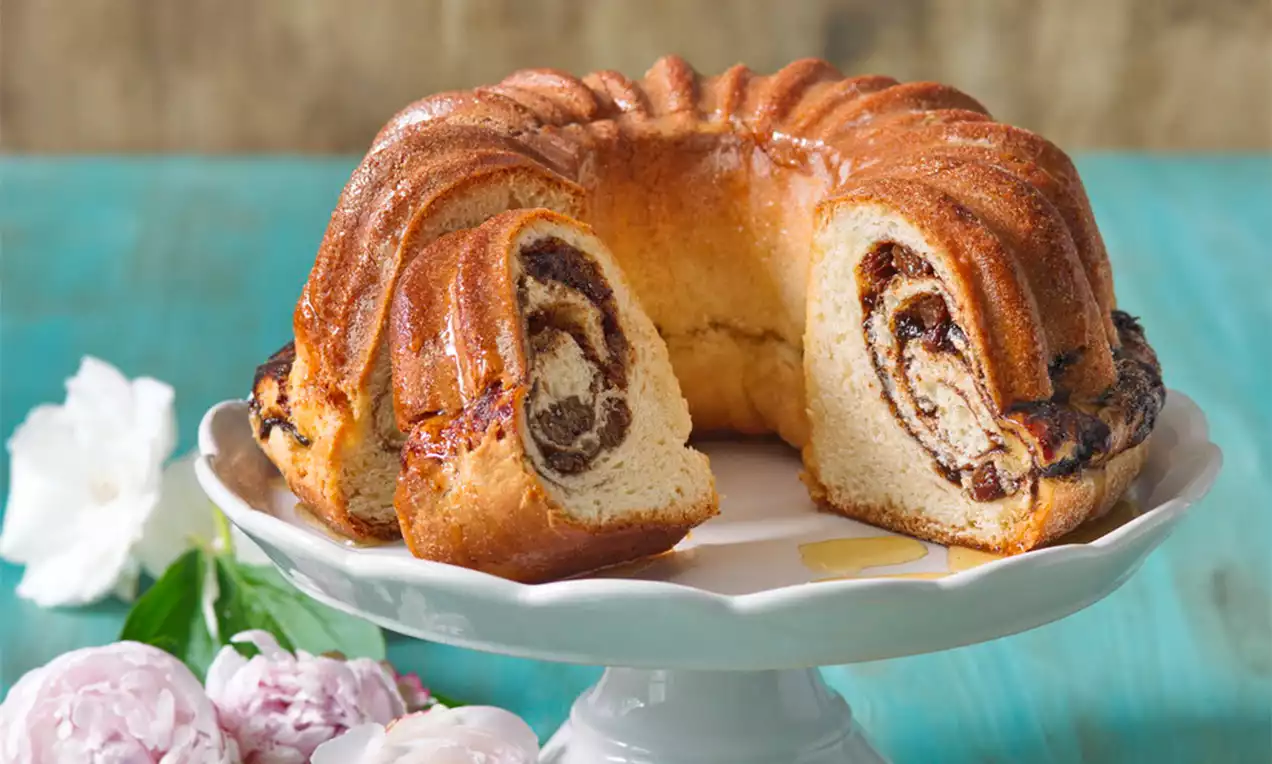 Kynutá bábovka zlatohnědé barvy s lesklým povrchem, uvnitř spirály plněné tmavou náplní, na bílé dortové míse, vedle růžových květů