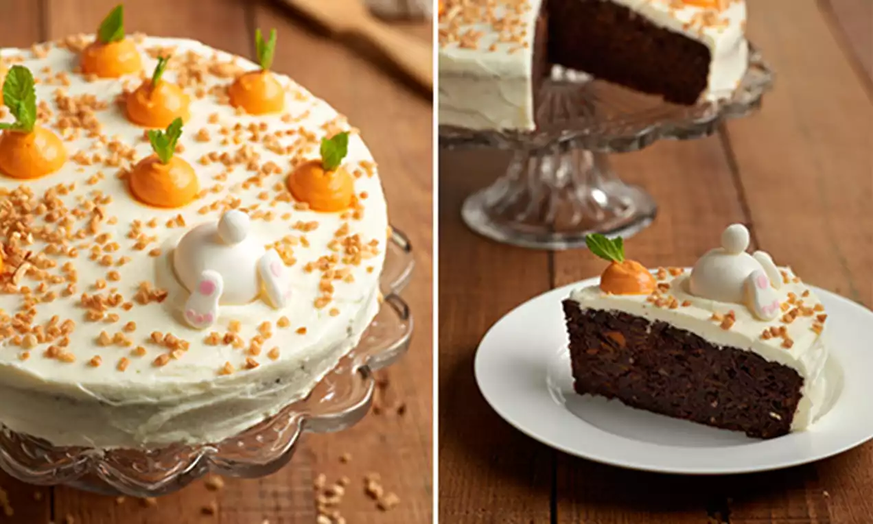 Tarta de bizcocho oscuro, cubierta de crema blanca, decoración de zanahorias naranjas con hojas verdes y migas crujientes sobre plato transparente