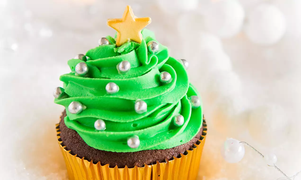 Cupcake avec un glaçage vert vif en spirale imitant un sapin, décoré de perles argentées et surmonté d'une étoile dorée sur fond neigeux