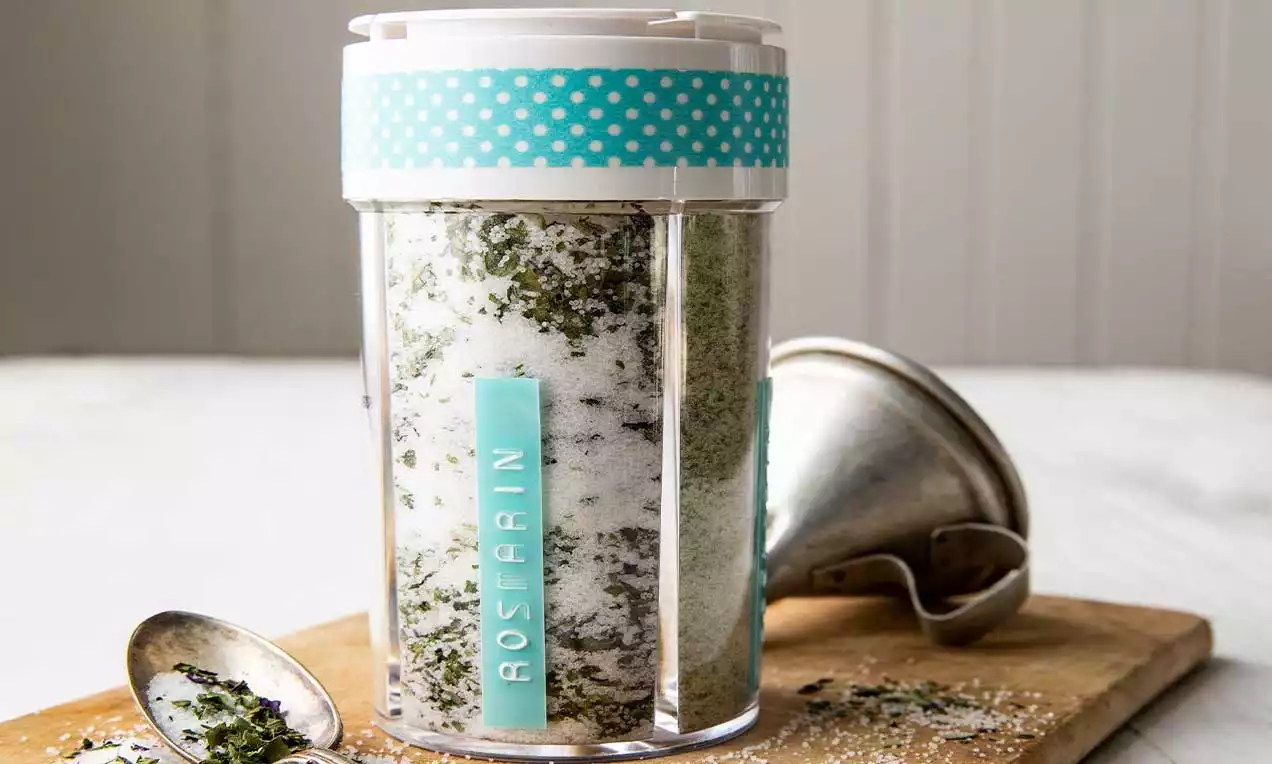 Feines weißes Salz und dunkelgrüne Kräuter, geschichtet in einem transparenten Glas mit türkisfarbenem Deckel, auf Holzbrett mit Löffel und Trichter