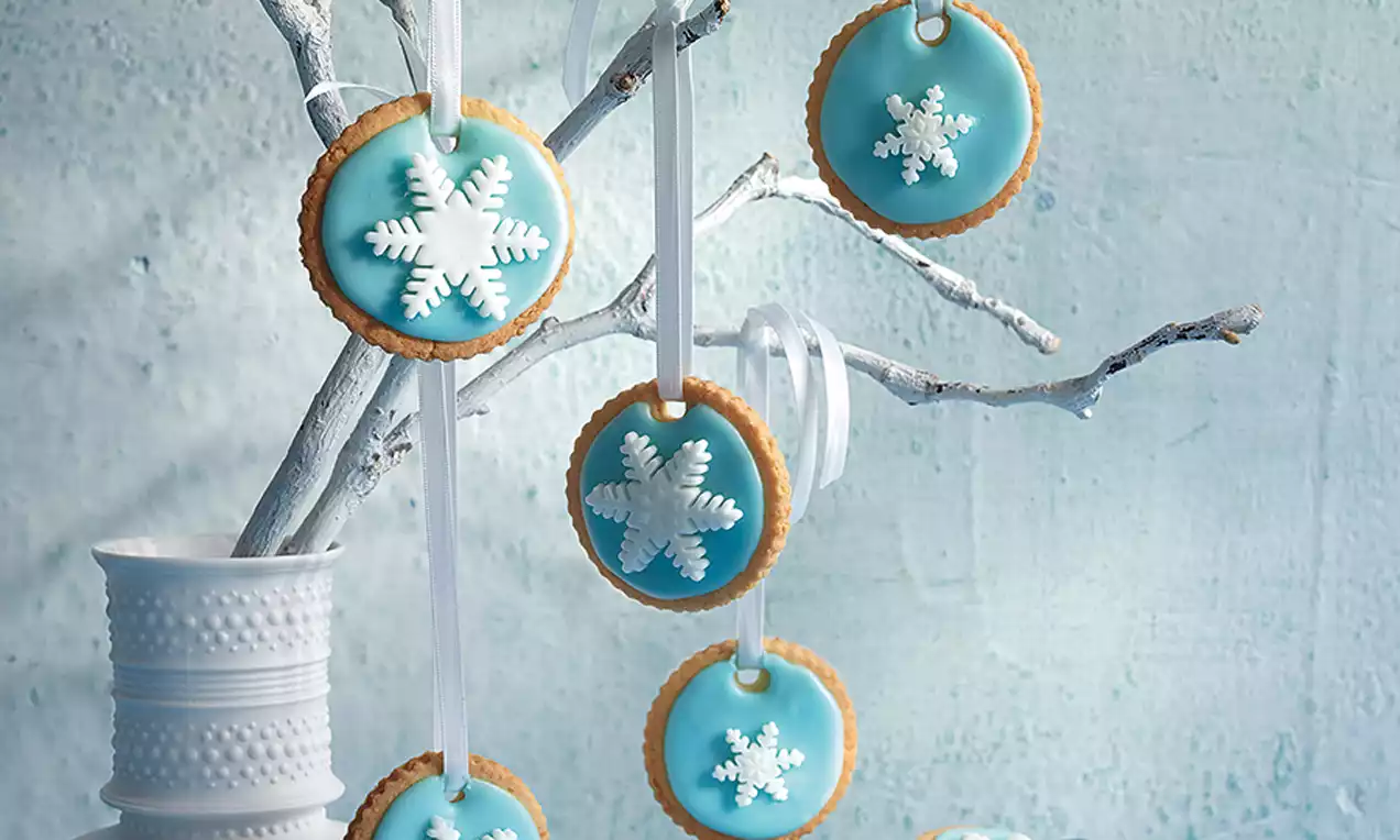 Runde småkaker med lys blå glasur og hvite snøfnugg av pynt hengt opp med hvite bånd på en hvitmalt gren i vase