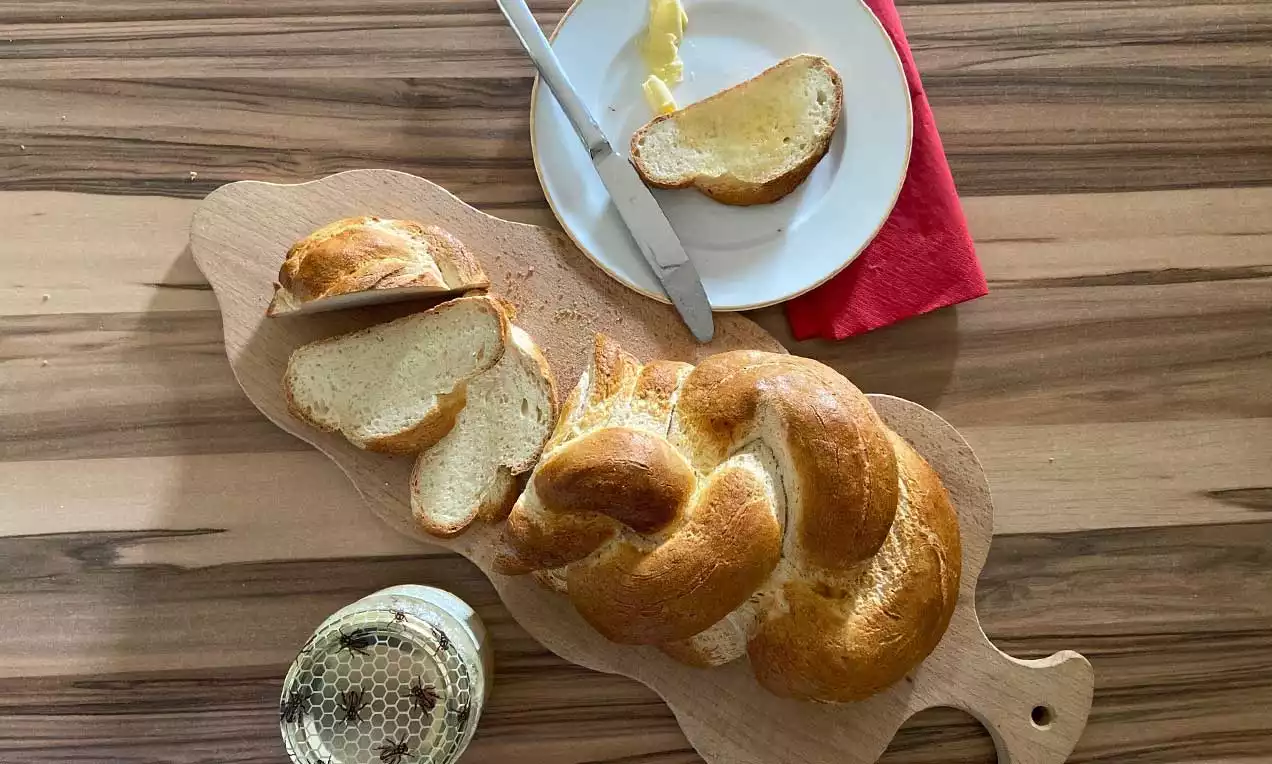 Goldbrauner glutenfreier Butterzopf mit glänzender Kruste, angeschnitten auf einem Holzbrett, daneben Butter und Brot auf weißem Teller