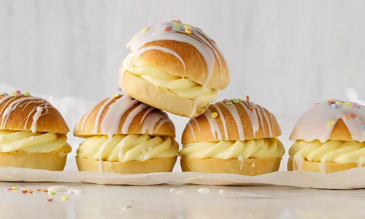 Fünf goldbraune Berliner mit glänzendem Zuckerguss und bunten Streuseln, gefüllt mit geschwungener gelber Vanillecrème auf hellem Papier