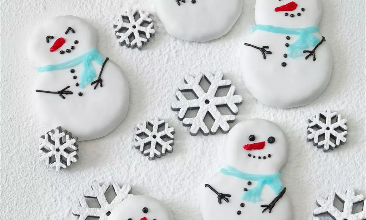 Biele snehuliaky z jemného cesta s tenkou lesklou polevou, zdobené modrými šálmi a čiernymi detailmi, rozložené s trblietavými vločkami na bielom povrchu