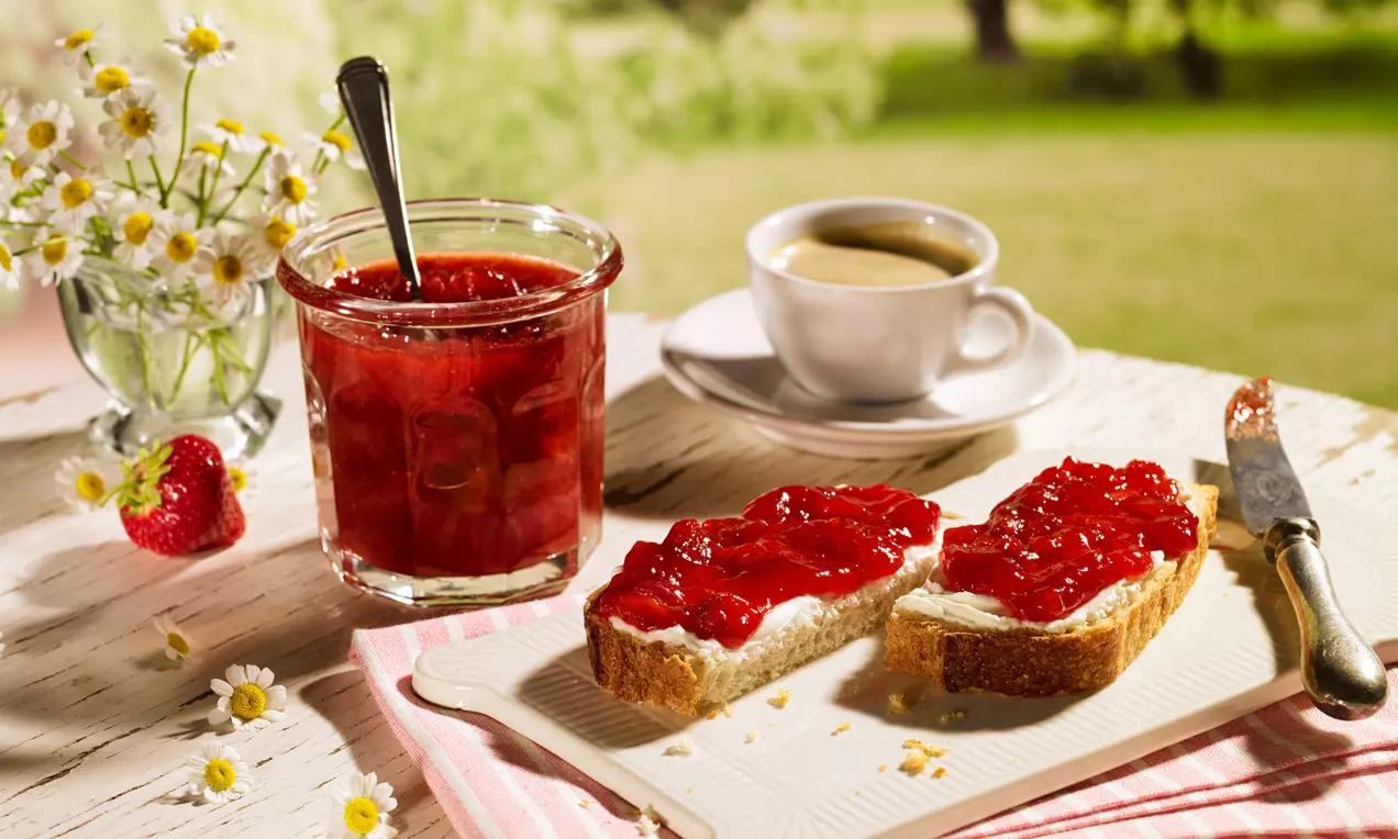 Pohár sýtočerveného jahodového džemu so lyžičkou vedľa krajcov chleba natretých maslom a džemom, na svetlom drevenom stole s kvetmi a kávou