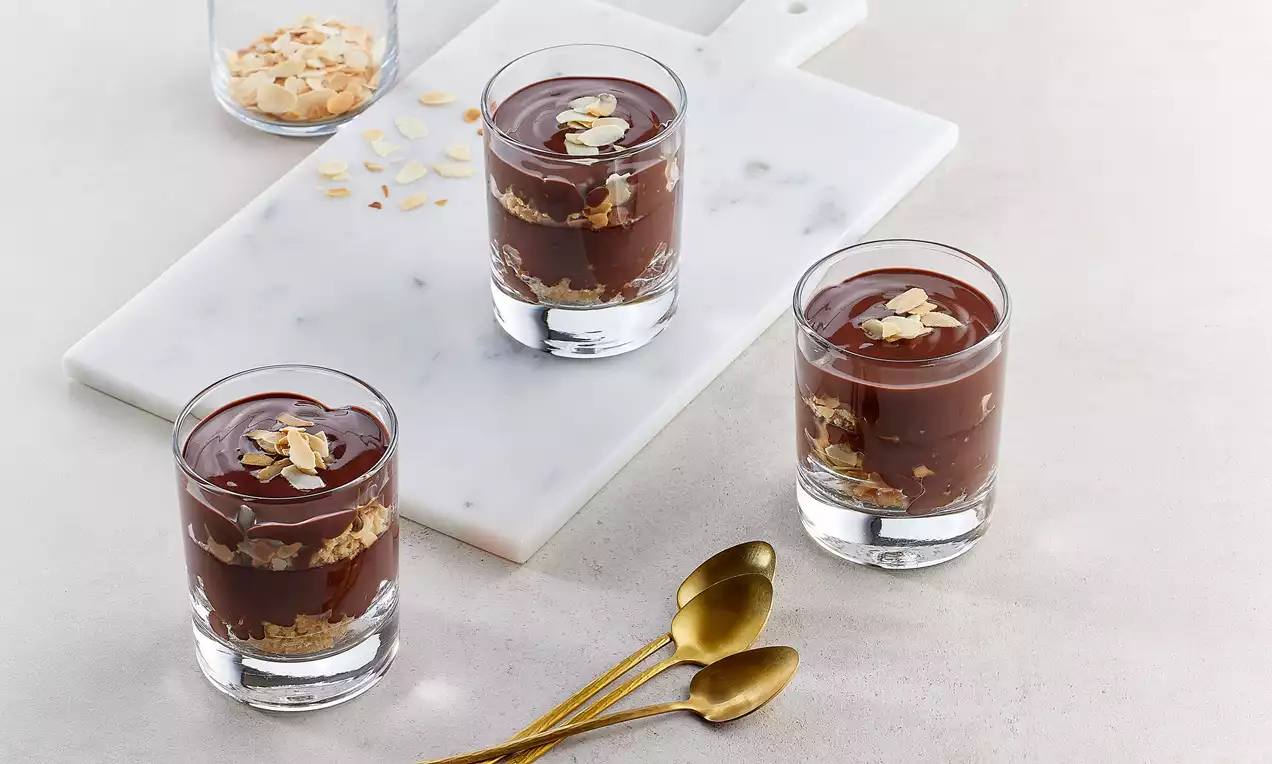 Cam kupa bardaklarda sunulan bademli bitter çikolatalı puding, altın kaşıklarla eşlik eden badem dilimleriyle zarif bir şekilde süslenmiş