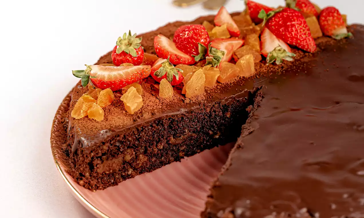 Tartă de ciocolată cu glazură lucioasă, decorată cu căpșuni roșii și bucăți de caise aurii pe un platou roz